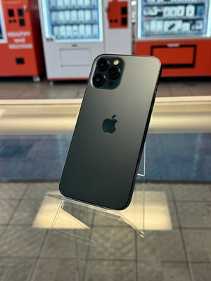 A dark gray Apple iPhone shown from the back, resting on a clear acrylic display stand in a retail or showroom setting with vending machines and tiled walls visible in the background.