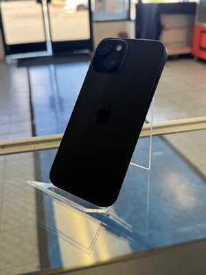 A black Apple iPhone shown from the back, resting on a clear acrylic display stand inside what appears to be a store or showroom.