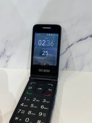 An Alcatel flip phone with a black exterior and a blue digital display screen, resting on a white marble-patterned surface.