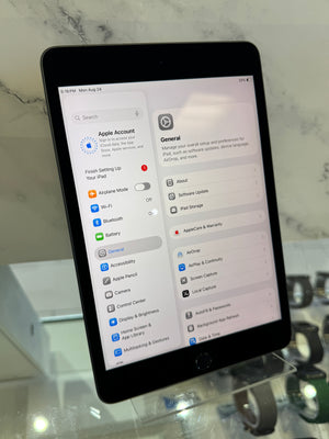 A black Apple iPad standing upright on a clear display stand, showing the iPad General settings screen on its screen. The background is a light marble-patterned surface.