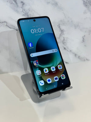 A black smartphone with a front-facing camera and a colorful home screen, displayed upright on a small gray stand against a white marble-patterned background.