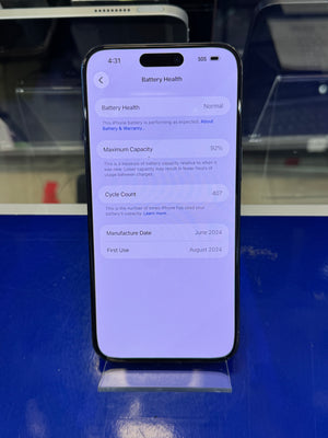 A black smartphone displayed upright on a clear stand, showing the battery health screen with 92% capacity and a 4:31 time indicator. The phone is photographed against a blue display surface with blurred store fixtures in the background.