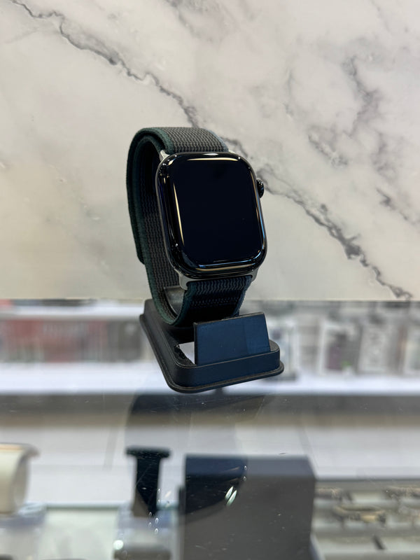 A smartwatch with a dark green mesh band and black screen, resting on a black charging dock on a glass counter with a marble-patterned background.
