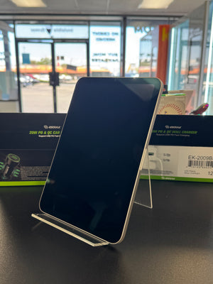 A black smartphone with a silver edge, resting on a clear acrylic stand on a dark tabletop. A green and black box is visible in the background, with a blurred storefront window behind it.