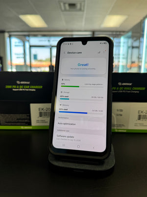 A smartphone on a charging stand in a retail or office environment, with two black and green Genuk brand boxes visible in the blurred background.