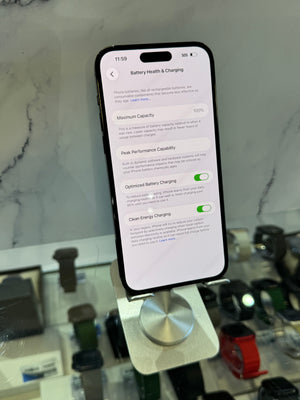 A smartphone on a display stand showing the Settings app page for Battery Health & Charging, photographed in a retail or showroom environment with blurred watches and a marble-patterned wall in the background.