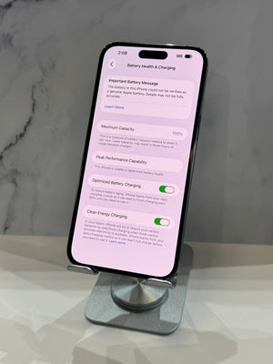 A black smartphone displayed upright on a silver stand, showing the Settings screen for Battery Health & Charging on a light pink background. The phone is photographed indoors against a light gray marble-like surface and wall.