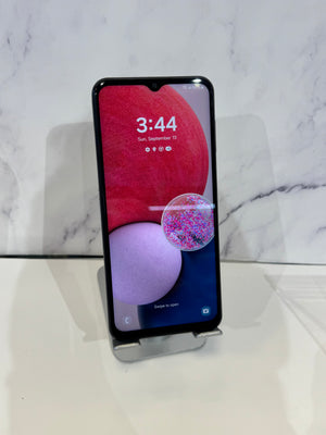 A black smartphone with a front-facing camera cutout stands upright on a small clear display stand against a white marble-patterned background.