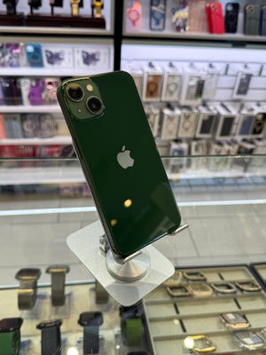 A dark green Apple iPhone 12 Pro shown from the back, mounted on a display stand inside a retail store with glass cases and accessories visible in the background.