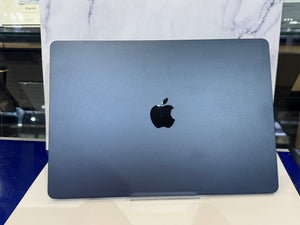 A closed Apple laptop in a dark blue-gray finish, standing upright on a white surface with a marble-patterned backdrop behind it.