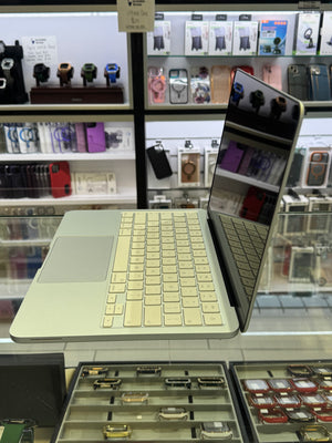A silver Apple MacBook laptop with a cream-colored Magic Keyboard is shown open on a retail display counter inside a store. The background features shelves stocked with various phone accessories, watch bands, and other gadgets.