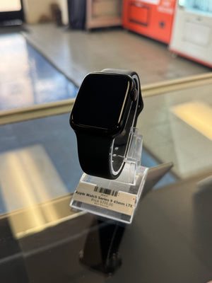 A black smartwatch with a square screen and black band, attached to a clear plastic retail tag, photographed on a glass display counter.