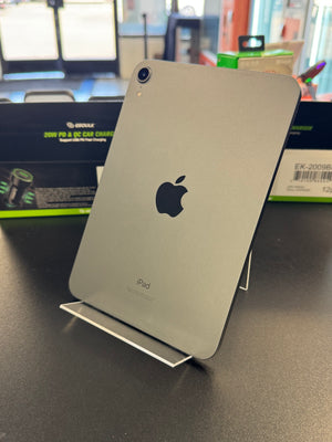 A silver Apple iPad shown from the back, resting on a clear display stand on a dark tabletop with store packaging visible in the blurred background.