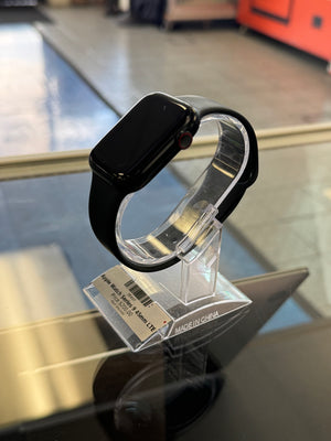 A black smartwatch with a black band displayed on a clear plastic stand, with a price tag attached. The watch is photographed inside what appears to be a store or display setting, with a blurred background.