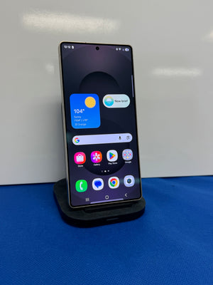 A black smartphone displayed upright on a small black stand against a white wall, with a blue fabric surface below. The phone screen is on and showing the Google home interface with app icons.