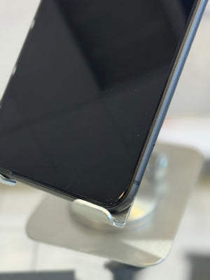 Close-up of a black smartphone resting on a metallic display stand, showing the top edge and front bezel against a softly blurred light background.