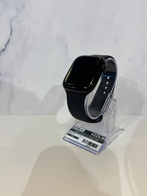 Black smartwatch with a glossy finish and black sport band, displayed on a clear acrylic stand with a price tag in front, set against a white marble-patterned background.