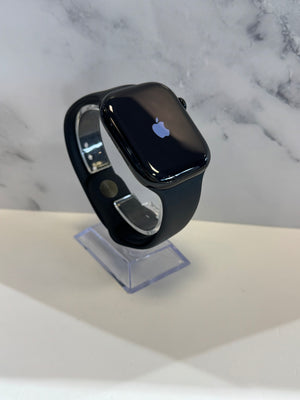 A black Apple Watch with a black sport band, displayed on a clear acrylic stand against a white and gray marble-patterned background.