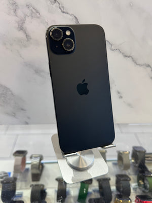 A black Apple iPhone 12 Pro Max is shown from the back, resting on a white display stand on a white marble-patterned surface. The Apple logo is visible in the center, and a watch display case is visible in the blurred background on the right.