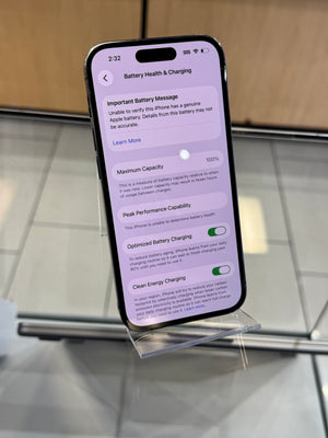 A smartphone displaying battery health and battery charging settings, photographed on a glass display stand in an indoor setting.