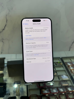 A black smartphone displaying the iPhone Battery Health settings screen, held upright in front of a marble-patterned background with a blurred retail display case below.