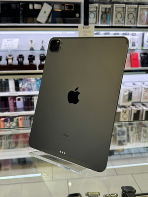 A dark gray Apple iPad Pro standing upright on a clear acrylic display stand inside a retail store, with shelves of phones and accessories blurred in the background.
