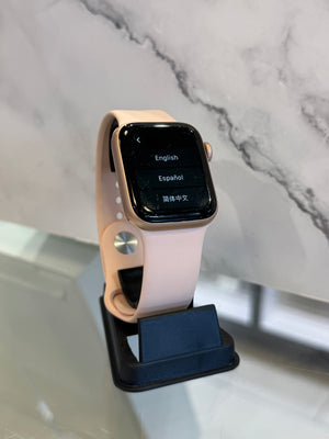 A smartwatch with a light pink band and a gold-toned case stands on a black display stand against a white marble-patterned background.
