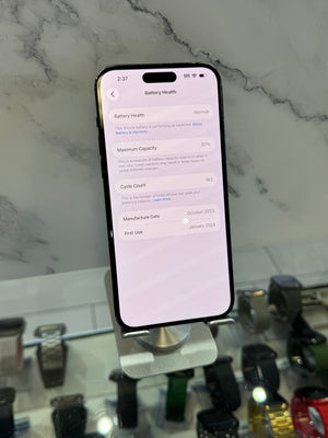 A black smartphone displayed upright on a stand, showing the battery health settings screen. The phone is photographed in a retail or display environment with marble-patterned wall and other watches visible in the blurred background.