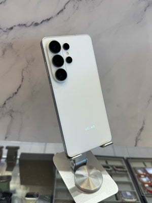 A white Samsung Galaxy S24 Ultra smartphone shown from the back, mounted on a silver display stand against a light marble-patterned wall.