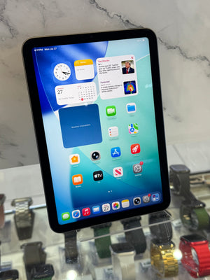 A black iPad Pro tablet on a clear display stand, showing the home screen with various app icons. The tablet is photographed in a retail or showroom setting with marble-patterned walls and other watch displays visible in the background.