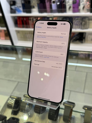 A black smartphone displaying battery health details on its screen, standing on a clear display stand inside a retail store with blurred shelves in the background.