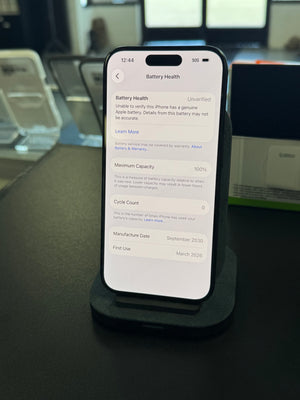 A smartphone displayed upright on a black stand on a dark tabletop, showing the Battery Health settings screen. The phone is photographed in a real indoor environment with blurred devices and equipment in the background.