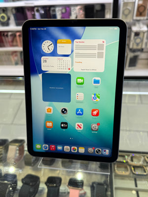A black iPad with its screen turned on, displaying the iOS home screen, standing on a clear display stand inside what appears to be a retail store.