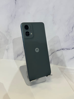 A dark gray Motorola smartphone shown from the back, resting on a small metallic display stand against a white marble-patterned background.