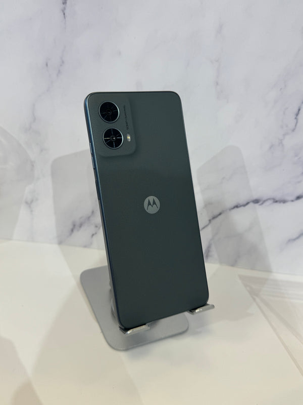 A dark gray Motorola smartphone shown from the back, resting on a small metallic display stand against a white marble-patterned background.