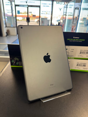 A silver Apple iPad shown from the back, standing upright on a small display stand on a dark tabletop. Apple's logo is visible in the center, with retail packaging boxes in the background inside a store.