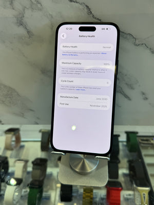 A black smartphone displayed upright on a metal stand, showing the battery health settings screen. The phone is photographed in a retail or showroom setting with a marble-patterned wall and watch displays blurred in the background.