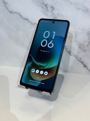 A black smartphone with a large front-facing screen displaying a blue and green lock screen, resting on a clear acrylic stand against a white marble-patterned background.