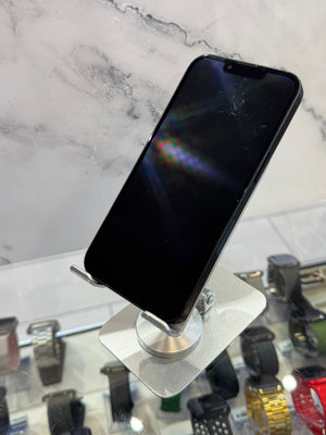 A black smartphone with a screen sits on a silver metal display stand in a retail setting, with a marble-patterned wall behind it and other watches or devices visible in the blurred background.