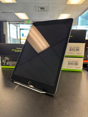 A black tablet device standing upright on a display stand on a dark tabletop, with product boxes visible in the background inside a retail or office environment.