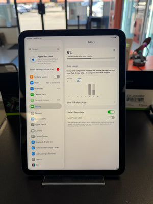 A black Apple iPad standing on a clear display stand on a dark tabletop, showing the Settings app screen with battery information. The background is a real indoor environment with blurred objects and windows.