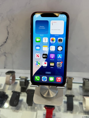 A black iPhone with a red bumper case stands on a silver metal display stand in a retail setting, with blurred smartwatches on a counter in the background.