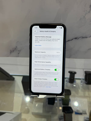 A black smartphone displayed upright on a stand, showing the Battery Health & Charging settings screen on its screen. The phone is photographed in a retail or showroom setting with a marble-patterned wall and other dark display items in the background.