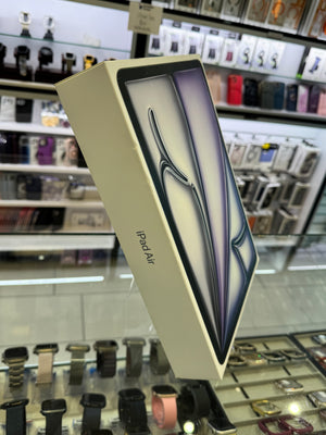 An iPad Air product box shown floating in front of blurred retail display cases containing watches and accessories. The box is cream-colored with a printed graphic of the tablet on top and the text 'iPad Air' visible on the side.