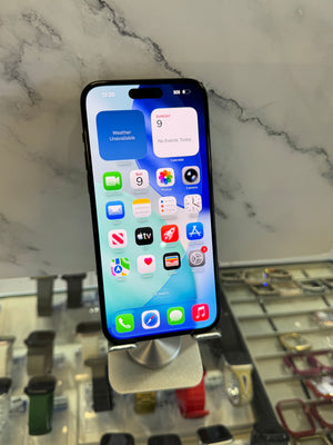 A black iPhone with its screen on, displaying the home screen, resting on a silver metal display stand in a retail setting with a marble-patterned background.