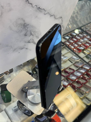 A black smartphone lying on its side on a small metallic display stand, photographed in a retail or showroom environment with shelves and fixtures blurred in the background.