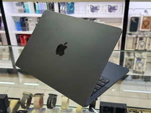 A closed Apple MacBook laptop in dark gray is shown at an angle on a glass display counter inside a retail store, with shelves of accessories blurred in the background.