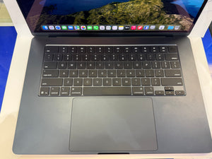A closed Apple MacBook laptop shown from a top-down angle, featuring a dark gray aluminum chassis, black keyboard with white lettering, and a large trackpad. The screen is turned on and displays the macOS desktop interface.