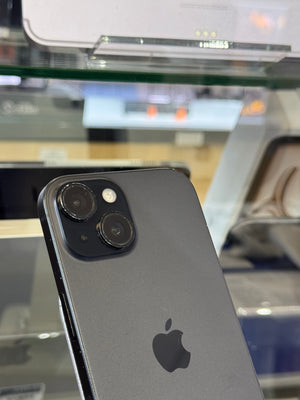 A close-up of the back of a dark gray Apple iPhone, showing the camera lenses and the Apple logo, photographed in a store or showroom setting with blurred glass cases in the background.