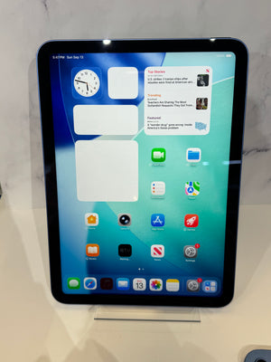 An iPad standing upright on a small display stand against a light marble-like background. The screen is on and shows the home screen with app icons and widgets.